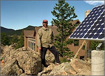 A photo of a man standing outside by a photovoltaic module installed on a rock outcropping with a house in background.
